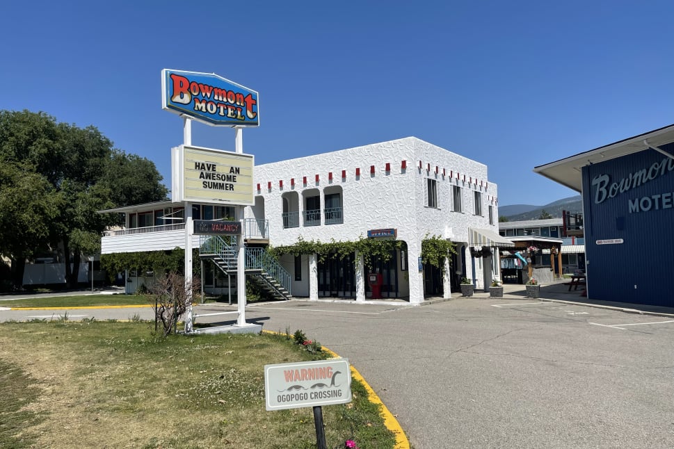A two-story white building of Bowmont Motel in Penticton with a "Have an awesome summer" sign, lush greenery, blue sky, and an OgoPogo crossing sign in the foreground.