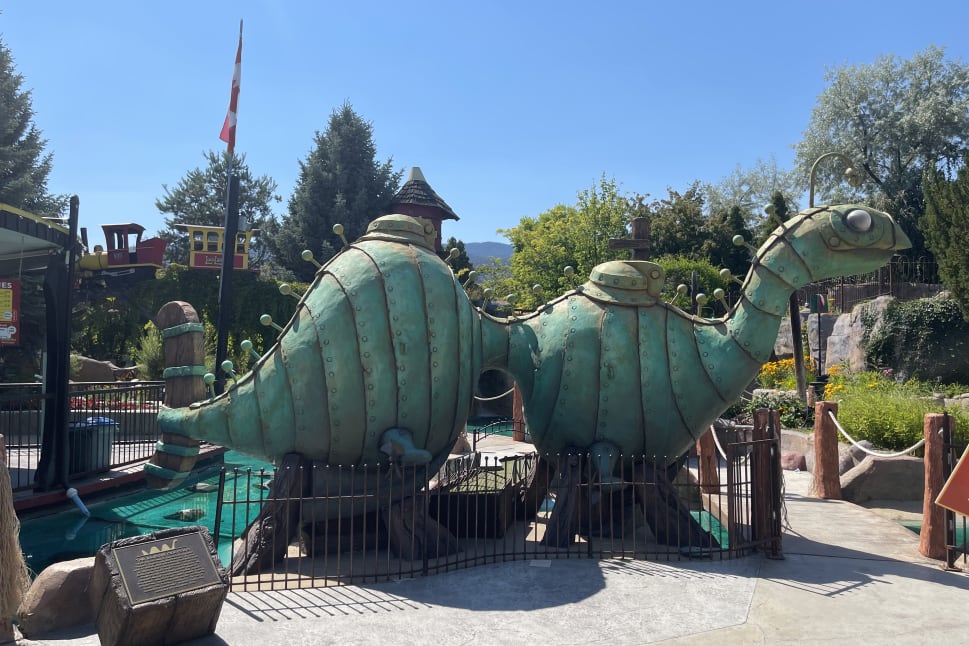 Large green dinosaur statue with bolts and knobs in an outdoor amusement park setting on a sunny day, Penticton.