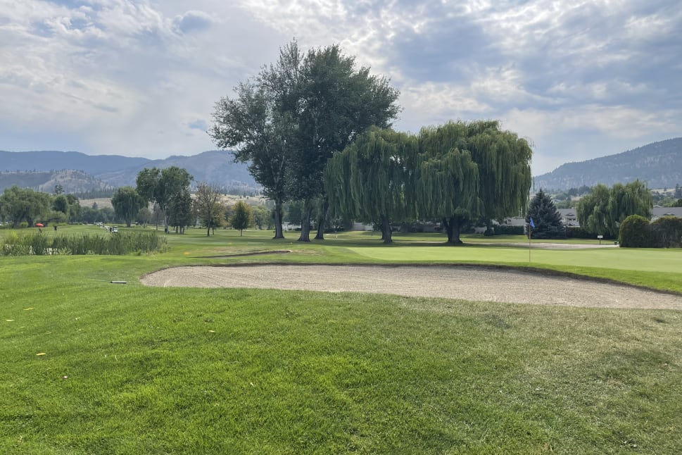 A picturesque golf course in Penticton, featuring lush green grass, a sand bunker, majestic trees, and mountainous backdrop under a partly cloudy sky.