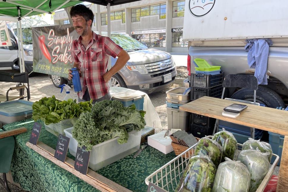 A vendor in a red checkered shirt stands behind a stall at a Kelowna farmers market, selling leafy greens like kale, chard, and lettuce, with a "Simply Salsa" sign in the background.