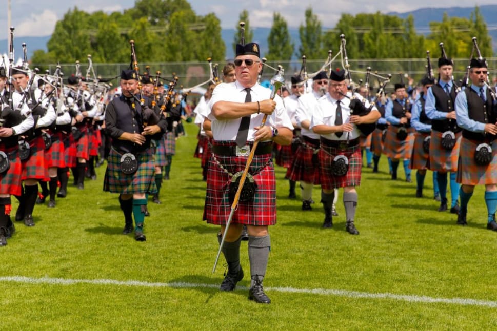 A group of bagpipers in traditional Scottish attire march on a grassy field during a Penticton event.