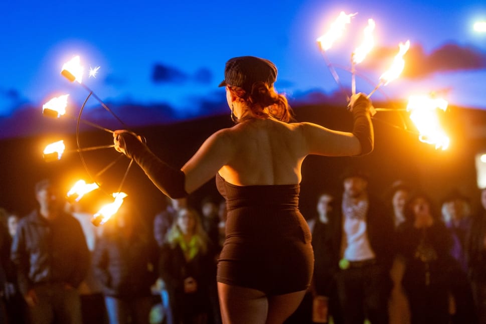 A performer with fiery sparklers entertains a nighttime crowd under a vibrant, colorful sky in Penticton.