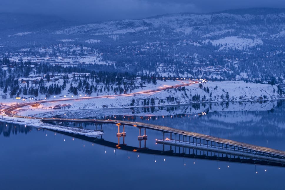 Aerial view of a snow-covered bridge and landscape in Kelowna, with lights reflecting on the calm water under a twilight sky.