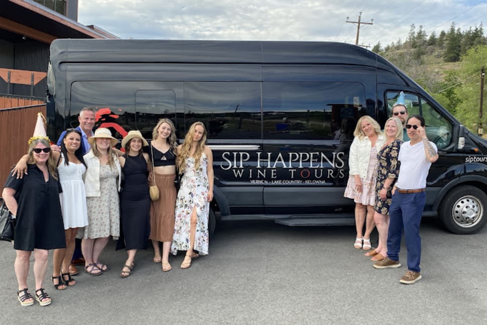 A group of people smiling and posing in front of a black van with "Sip Happens Wine Tours" in Vernon.