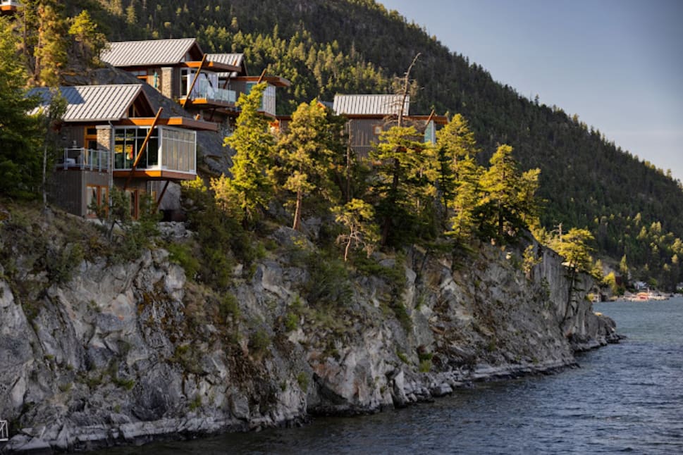 Houses perched on a rocky cliff overlooking a serene body of water in Vernon, surrounded by lush green trees and hills.