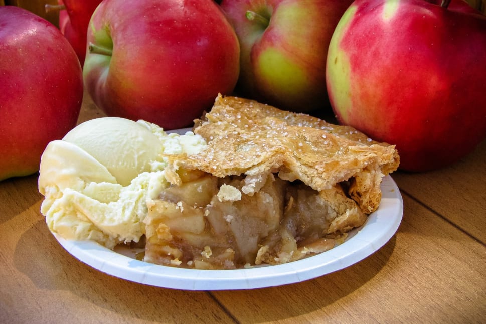 A plate of apple pie with a scoop of vanilla ice cream sits on a table surrounded by fresh red apples.