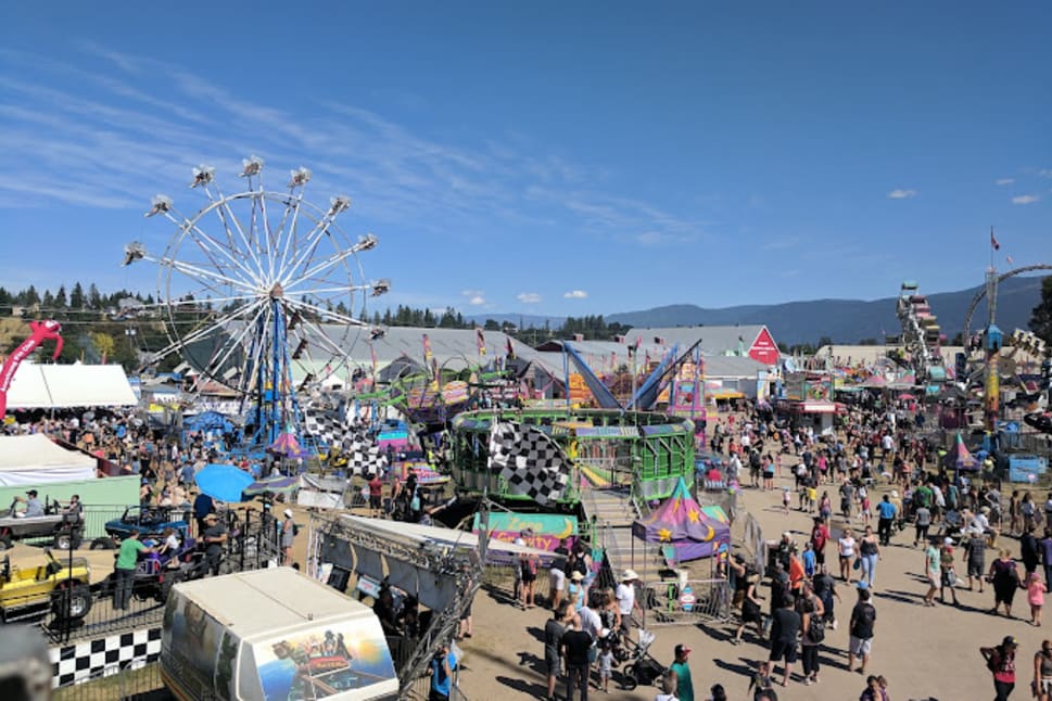 A busy fairground in Vernon with a Ferris wheel, colorful rides, and crowds of people enjoying a sunny day.