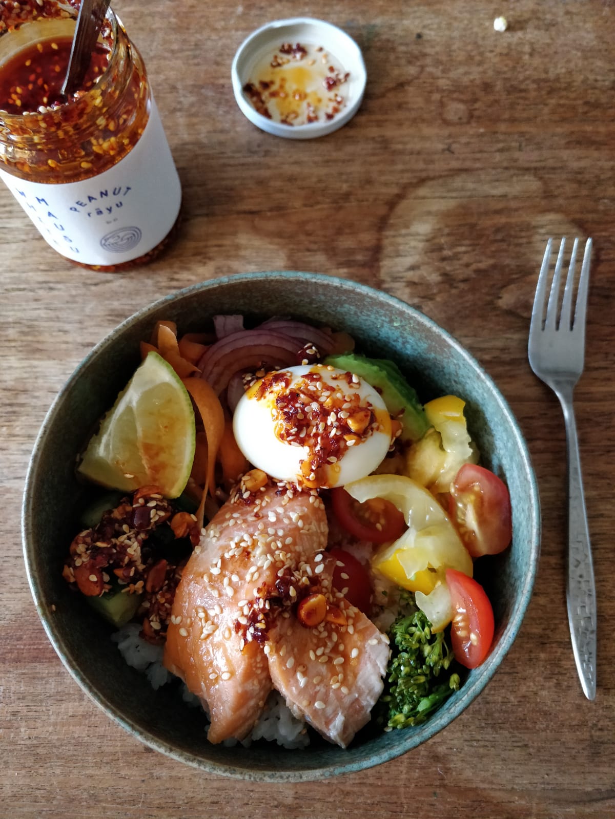 Poke bowl with boiled egg, salmon, lime wedge and varity of vegetables. A jar of Peanut Rayu stands on the table above it.