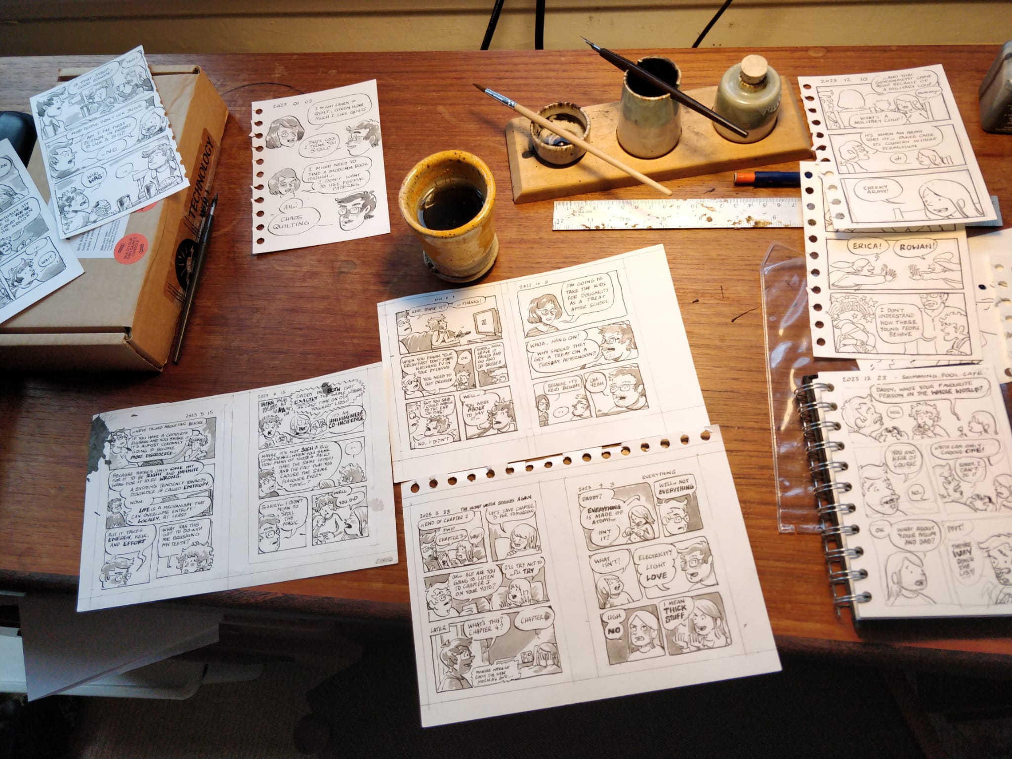 Multiple small comic strips in different stages of completion on a desk with ink pots, pencils, dip pens etc