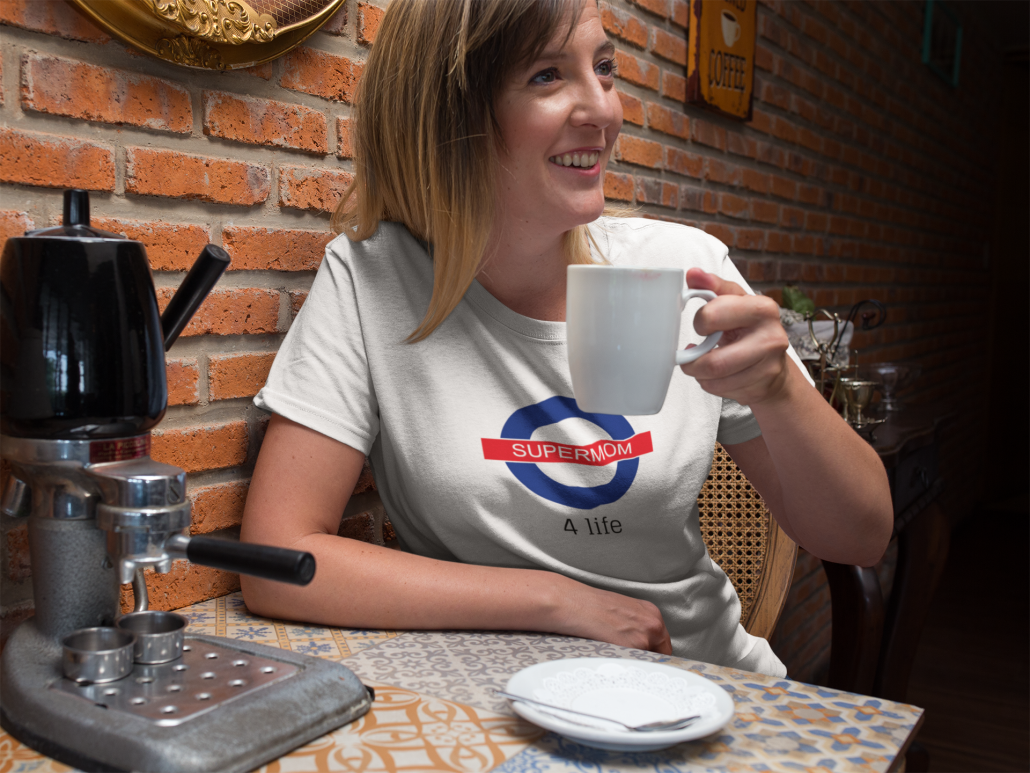 Frau in weißem T-Shirt mit Aufdruck "Supermom for life" sitzt am Tisch und trinkt Kaffee