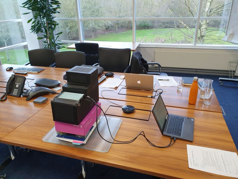 A conference table with various speaker equipment, a black, cardboard prototype, laptop and water jug