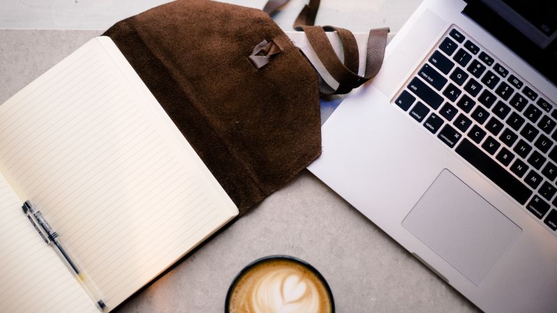 A notebook with pen, a MacBook and coffee on a table top