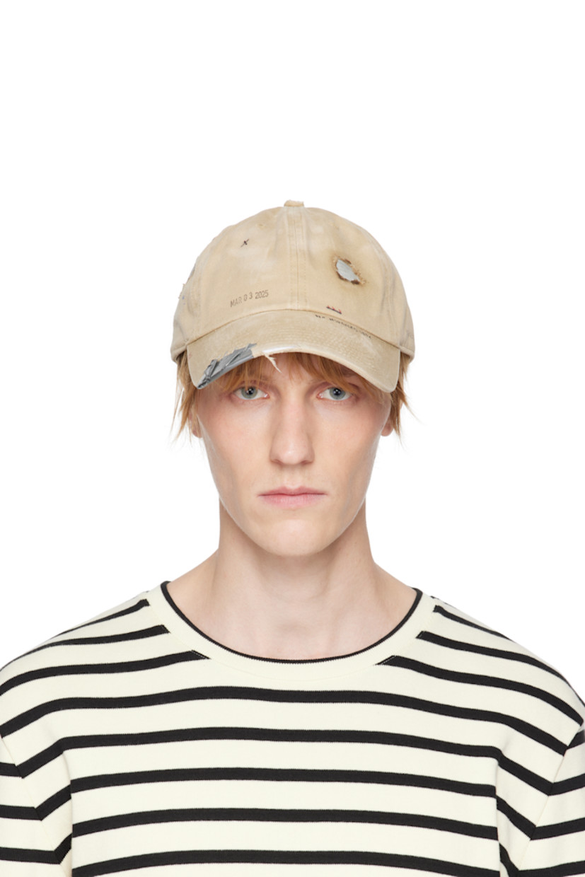 Cotton twill cap in tan. 

· Fading, distressing, and hand-drawn detailing throughout
· Embroidered eyelets at crown
· Text printed at face
· Staples and tape at curved brim
· Cinch fastening
· Plain-woven browband
· Tape at interior

Supplier color: Tan
