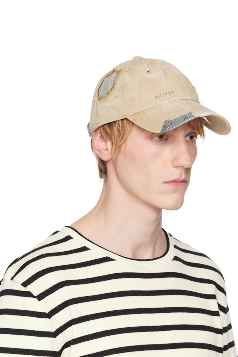 Cotton twill cap in tan. 

· Fading, distressing, and hand-drawn detailing throughout
· Embroidered eyelets at crown
· Text printed at face
· Staples and tape at curved brim
· Cinch fastening
· Plain-woven browband
· Tape at interior

Supplier color: Tan