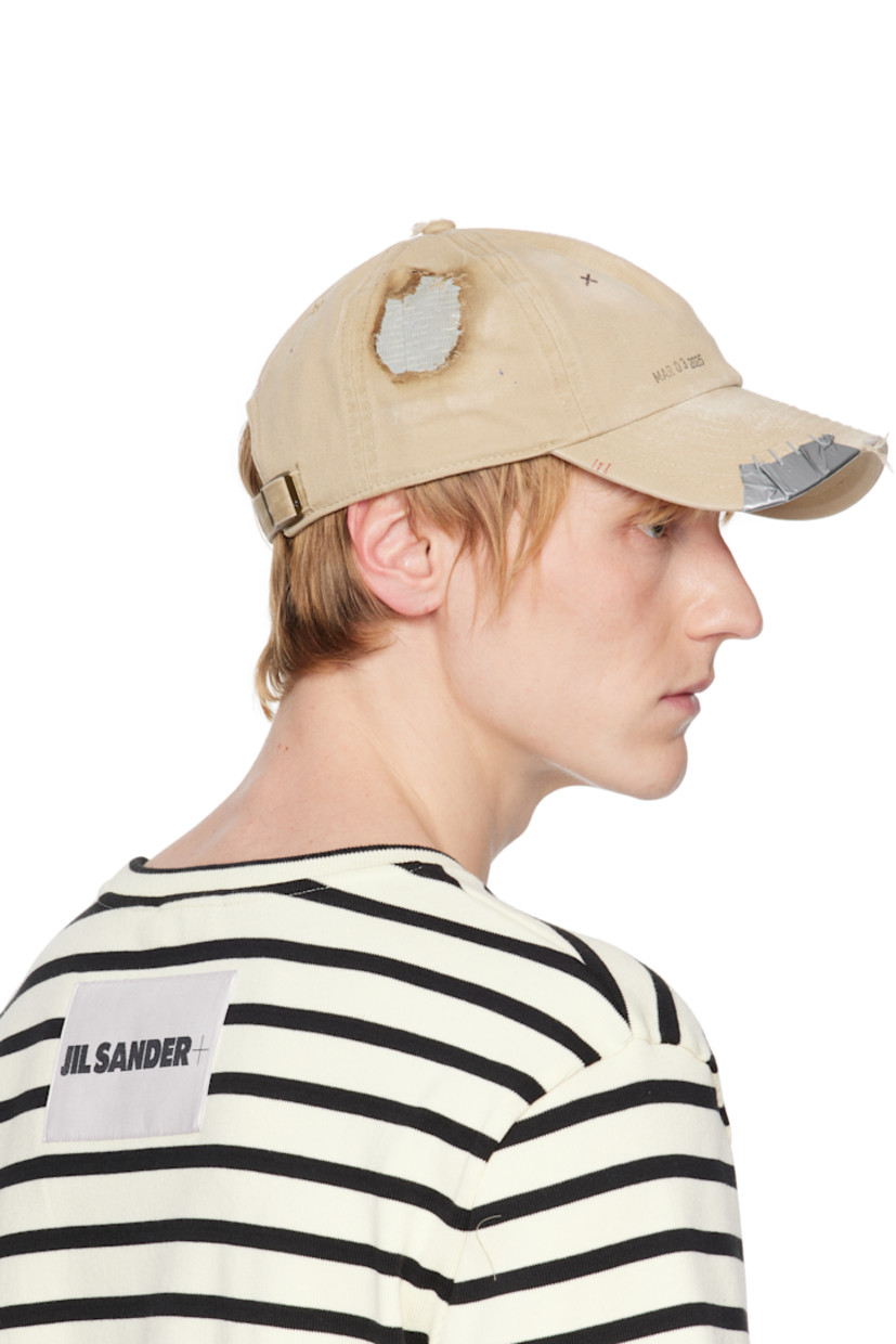 Cotton twill cap in tan. 

· Fading, distressing, and hand-drawn detailing throughout
· Embroidered eyelets at crown
· Text printed at face
· Staples and tape at curved brim
· Cinch fastening
· Plain-woven browband
· Tape at interior

Supplier color: Tan