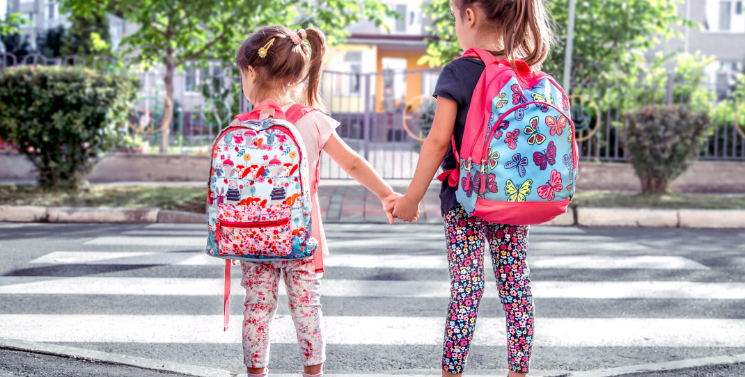 To jenter med ransel hånd i hånd ved fotgjengerfelt på vei til skolen