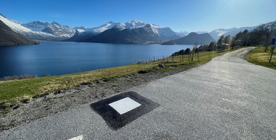 Fastmerke er et hvitt kvadrat inne i et svart kvadrat, malt direkte på asfaltvegen, i bakgrunn sees fjord og fjell.