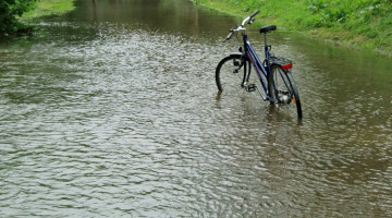 En sykkel står delvis nedsenket i flomvann i Storåna, der gangstien er oversvømt og vannet har bredt seg utover grøntområdene langs elva.