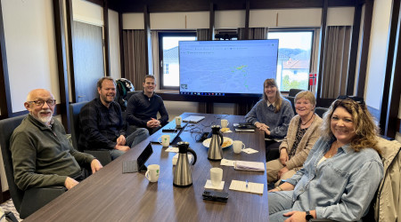 Seks personer smiler rundt et møtebord med kaffe og notater. Skjerm med kart vises i bakgrunnen