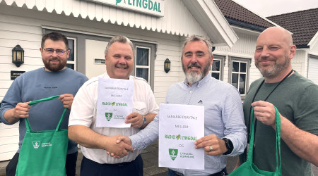 Bilde av fire personer foran bygget til Radio Lyngdal, de holder opp den signerte samarbeidsavtalen som er inngått mellom radioen og Lyngdal kommune