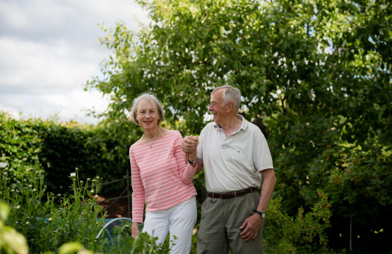 Eldre dame og mann som holder hender og smiler i en grønn og frodig hage.