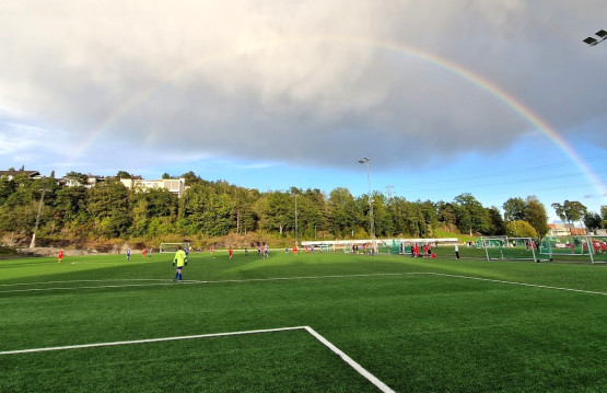 Fotballbane med spillere ute på banen. Regnskyer og regnbue i bakgrunnen.