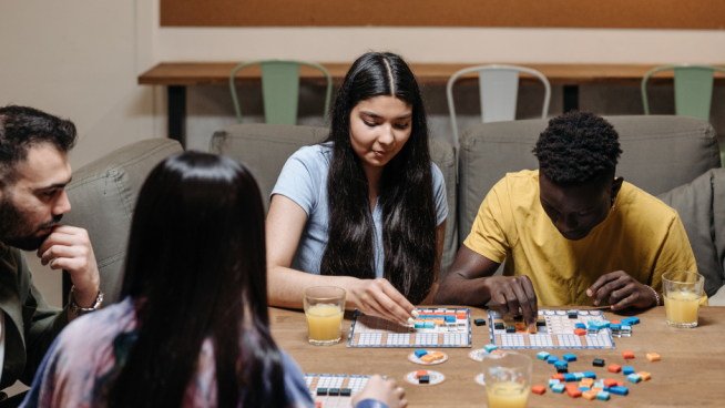 Fire personer sitter rundt et bord og spiller brettspill. På bordet står det fire glass med appelsinjuice og flere fargerike spillbrikker. I bakgrunnen henger en oppslagstavle med bilder og lapper.