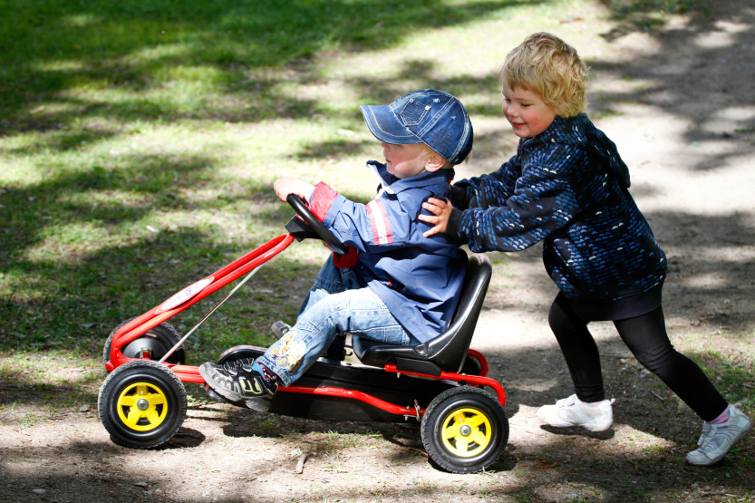 Et barn sitter i en tråbil og et barn står bak tråbilen og hjelper med å skape fart ved å dytte i ryggen.