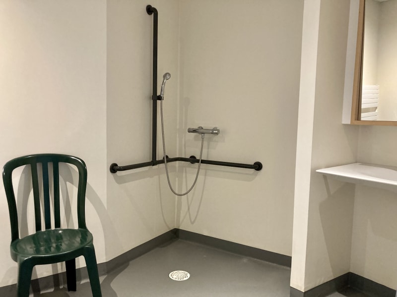 Une salle d'eau mono-bloc avec une douche à l'italienne, un lavabo et un large miroir, ainsi qu'une assise en plastique permettant une toilette dans une position confortable.