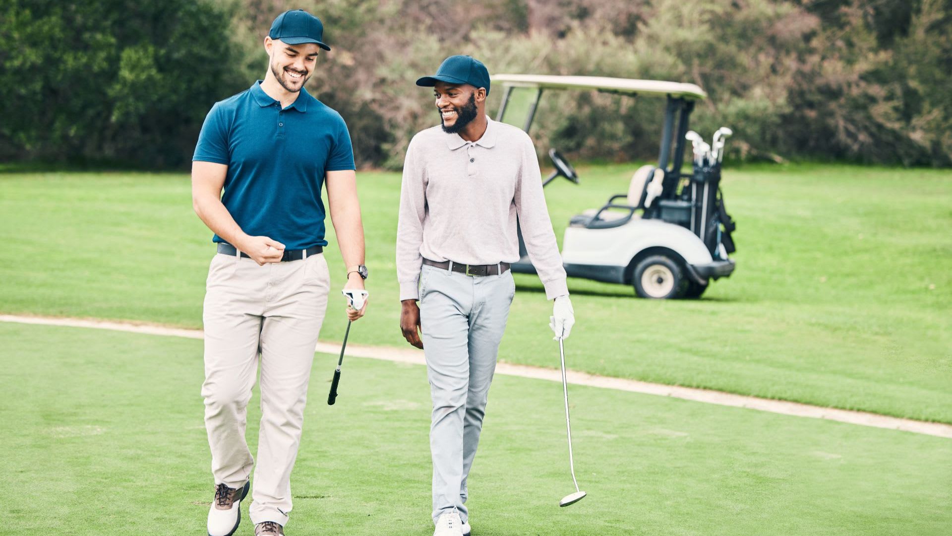Two men walking on a golf course, smiling and holding golf clubs from popular golf brands, with a golf cart and trees in the background.