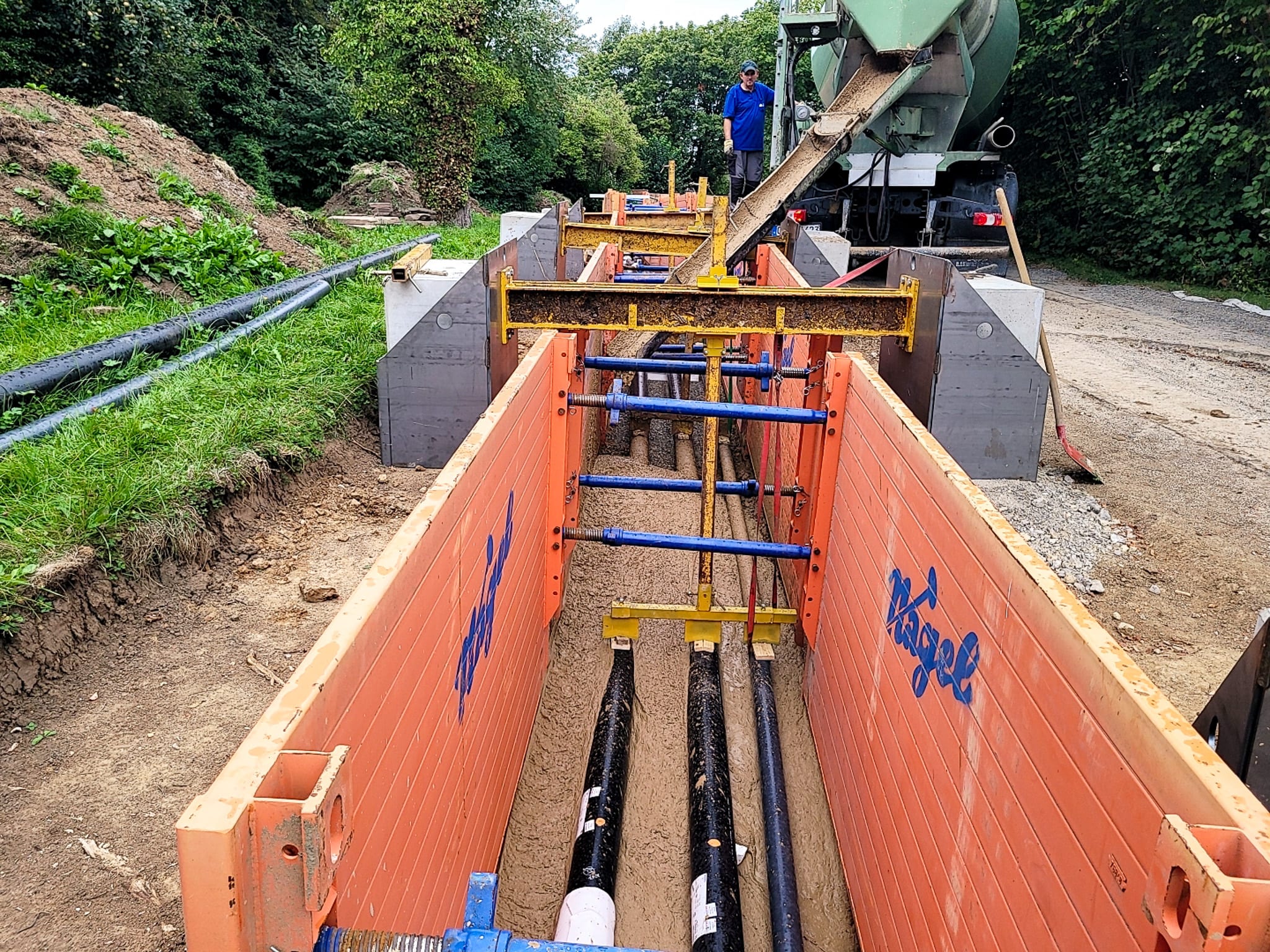 Innovative Fernwärmeerschließung mit Flüssigboden Ein tiefer Graben ist mit Wänden ausgeschachtet, am Boden des Grabens liegen, mehrere Rohre. Ein Zementmischer steht auf der Straße neben der Baustelle und füllt Flüssigboden in den Graben.