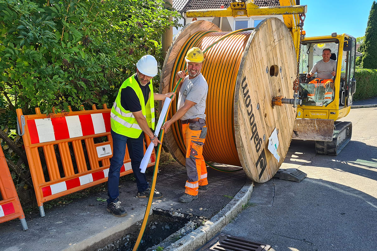 Ausbau Telekommunikation Maichingen Zwei Bauarbeiter verlegen ein Glasfaserkabel in einen geöffneten Schacht. Hinter ihnen steht die riesige Kabeltrommel. Ein dritter Bauarbeiter sitzt in einem Bagger.