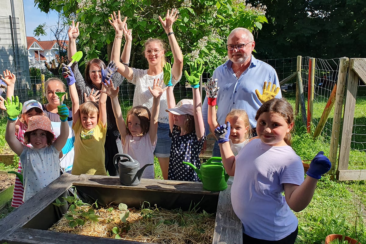 Gartenprojekt Kinderschutzbund Eine Gruppe Kinder und Herr Hoffmann sind im Schulgarten und stehen um ein Hochbeet herum. Die Kinder heben die Hände in die Luft und freuen sich.