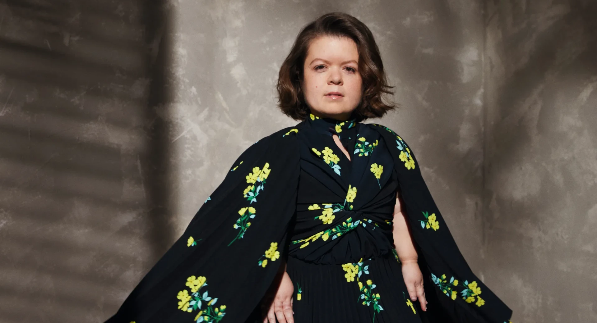 A person sitting in front of a textured wall with a black floral dress on. The floral print shows yellow flowers with turquoise leaves and green stems.