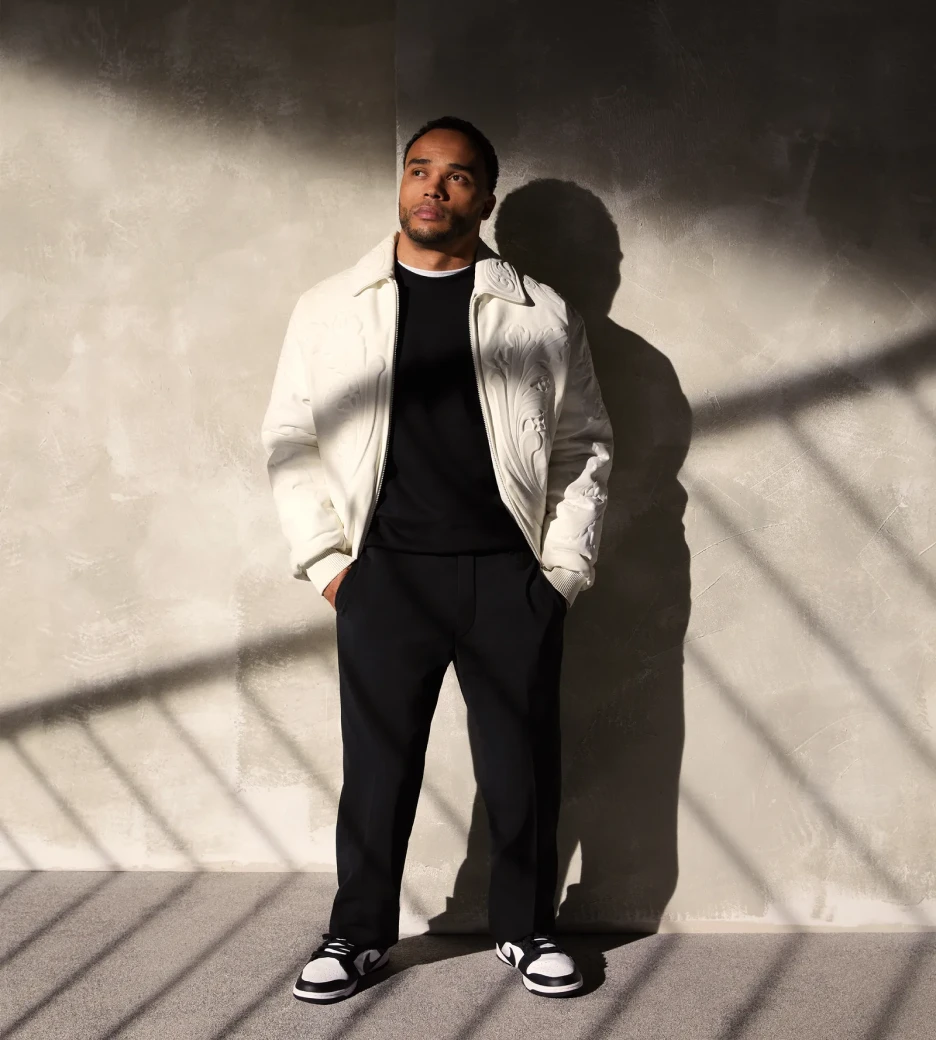 A person stands in front of a textuerd wall with stark lighting. They are wearing all black clothes except for white jacket.