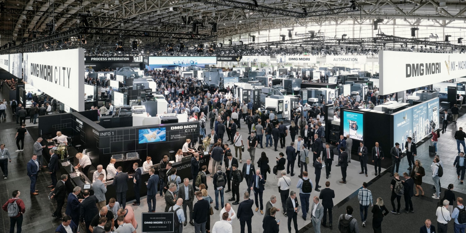 Große Messehalle mit dichtem Besucherandrang. Zahlreiche Stände, Maschinen und Displays unter dem Motto „DMG MORI CITY“. Helle Beschilderung und moderne Technik prägen die industrielle Ausstellung.