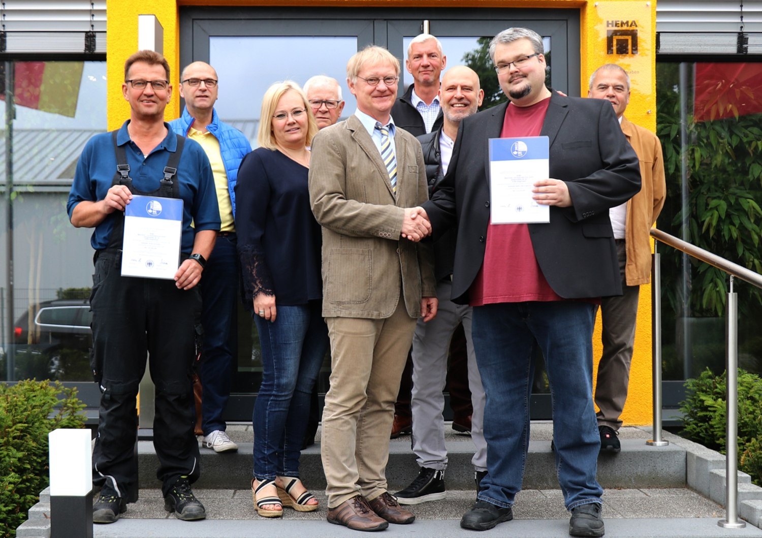 Das Team von Hema erhält die Urkunde zum BGHM-Gütesiegel ›Sicher mit System‹, die stellvertretend für den Gesamtbetrieb Sicherheitsfachkraft Sebastian Becker (rechts mit Urkunde) von Herwig Kochan zuständige BGHM-Aufsichtsperson (händeschüttelnd links) entgegennimmt (© HEMA Maschinen- und Apparateschutz GmbH) 