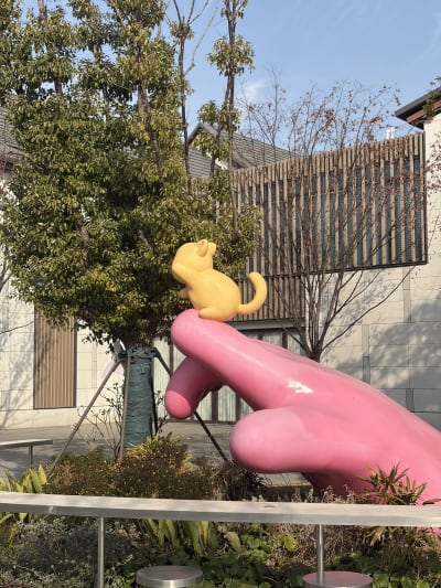 A large pink hand sculpture features a small yellow cat-like figure perched on its extended index finger. The installation is set outdoors in front of a leafy green tree and a modern building under a clear blue sky.