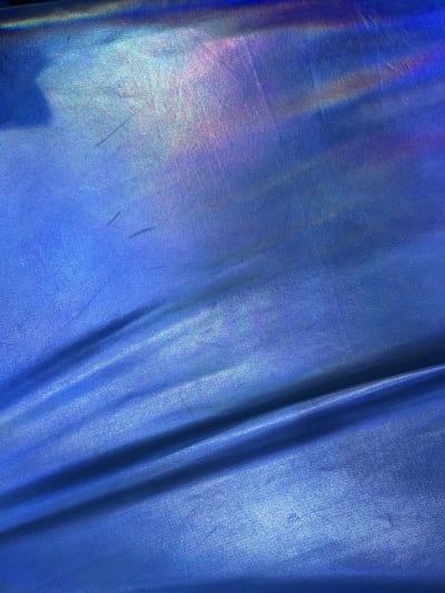 A shimmering, iridescent blue fabric with a fine vertical-ribbed texture and subtle rainbow-colored light reflections along the top edge. The material is draped with a soft horizontal fold near the bottom, creating variations in color and light.