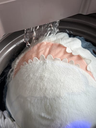Blahaj being washed in a washing machine, water pulling out from above, hitting his mouth