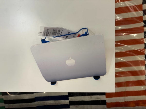 A silver MacBook prop sits on a blue LÅNESPELARE stand with a price tag of ¥19.9 on a white table. This top-down view also shows a striped rug beneath the table.
