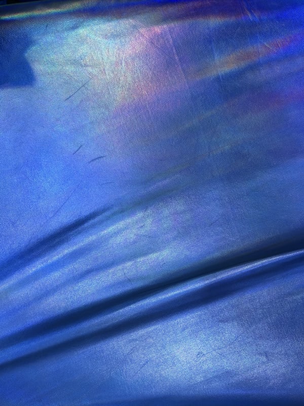 A shimmering, iridescent blue fabric with a fine vertical-ribbed texture and subtle rainbow-colored light reflections along the top edge. The material is draped with a soft horizontal fold near the bottom, creating variations in color and light.
