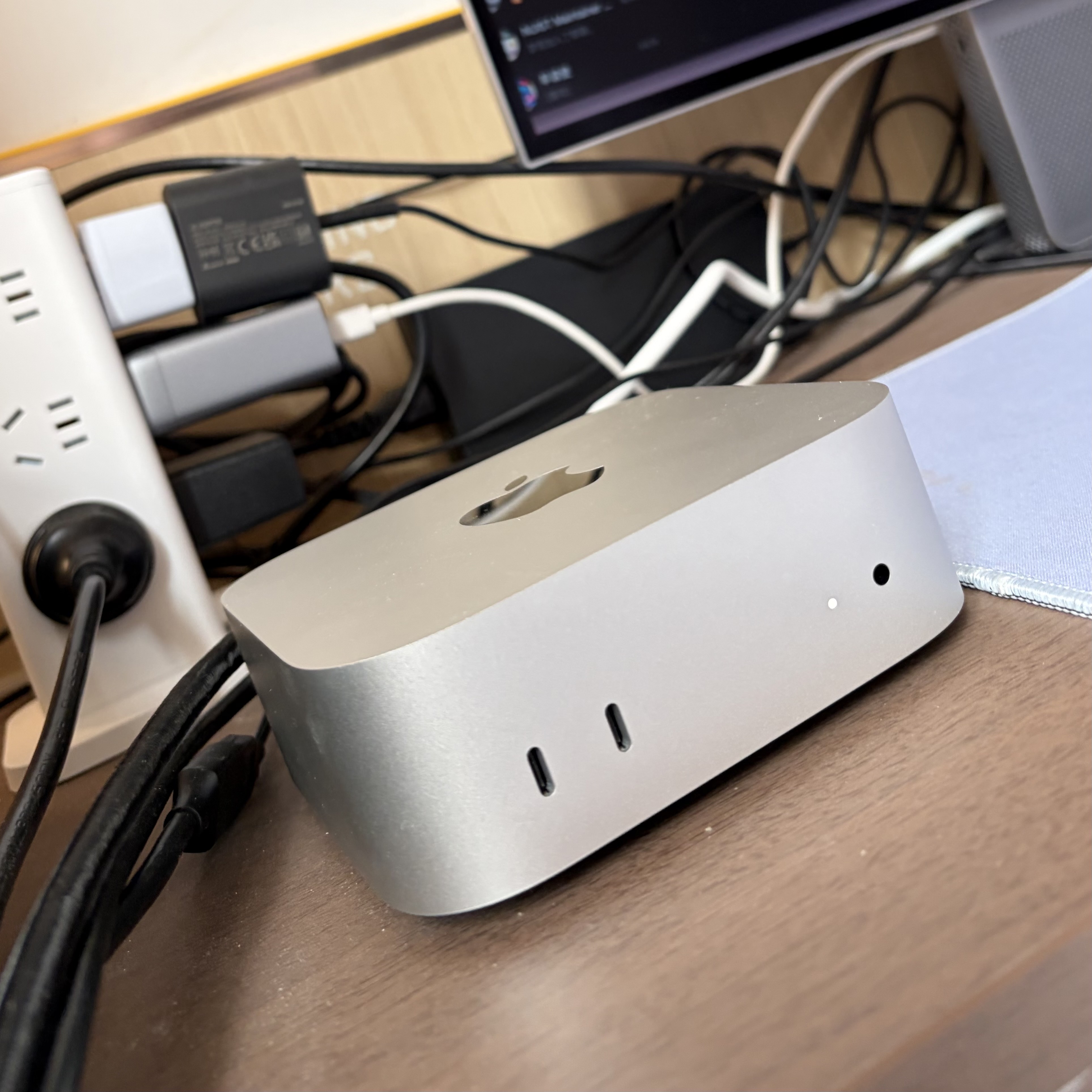 A close-up of a cluttered desk with tangled cables, power adapters, a power strip, and the base of a silver computer monitor.