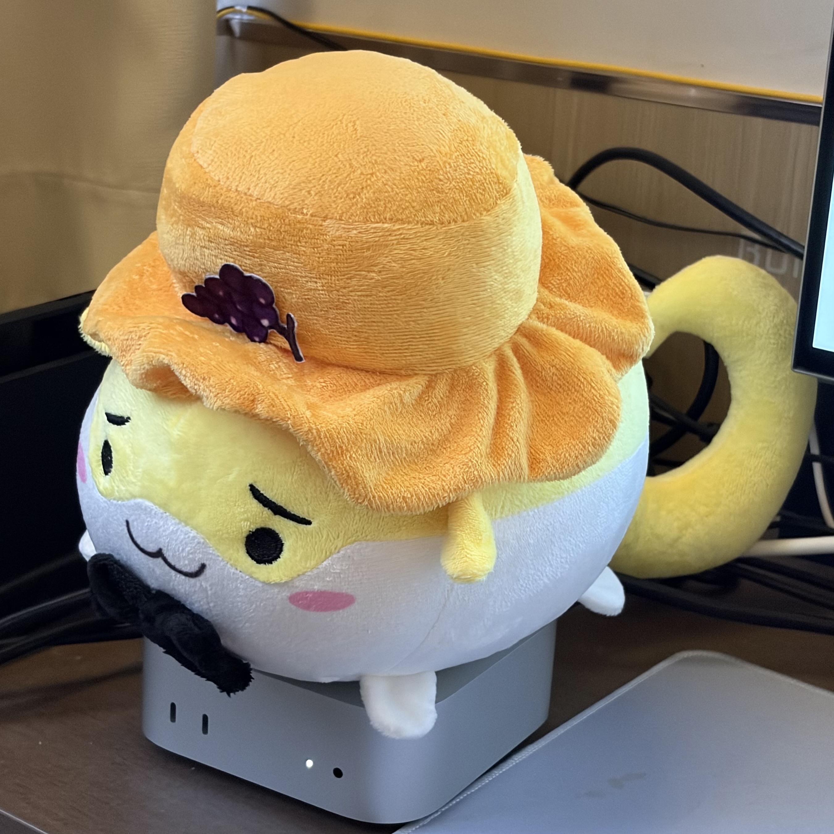 A round yellow-and-white plush character with a smiling face wears an oversized orange hat decorated with a purple grape cluster. It sits on a small gray device on a desk.