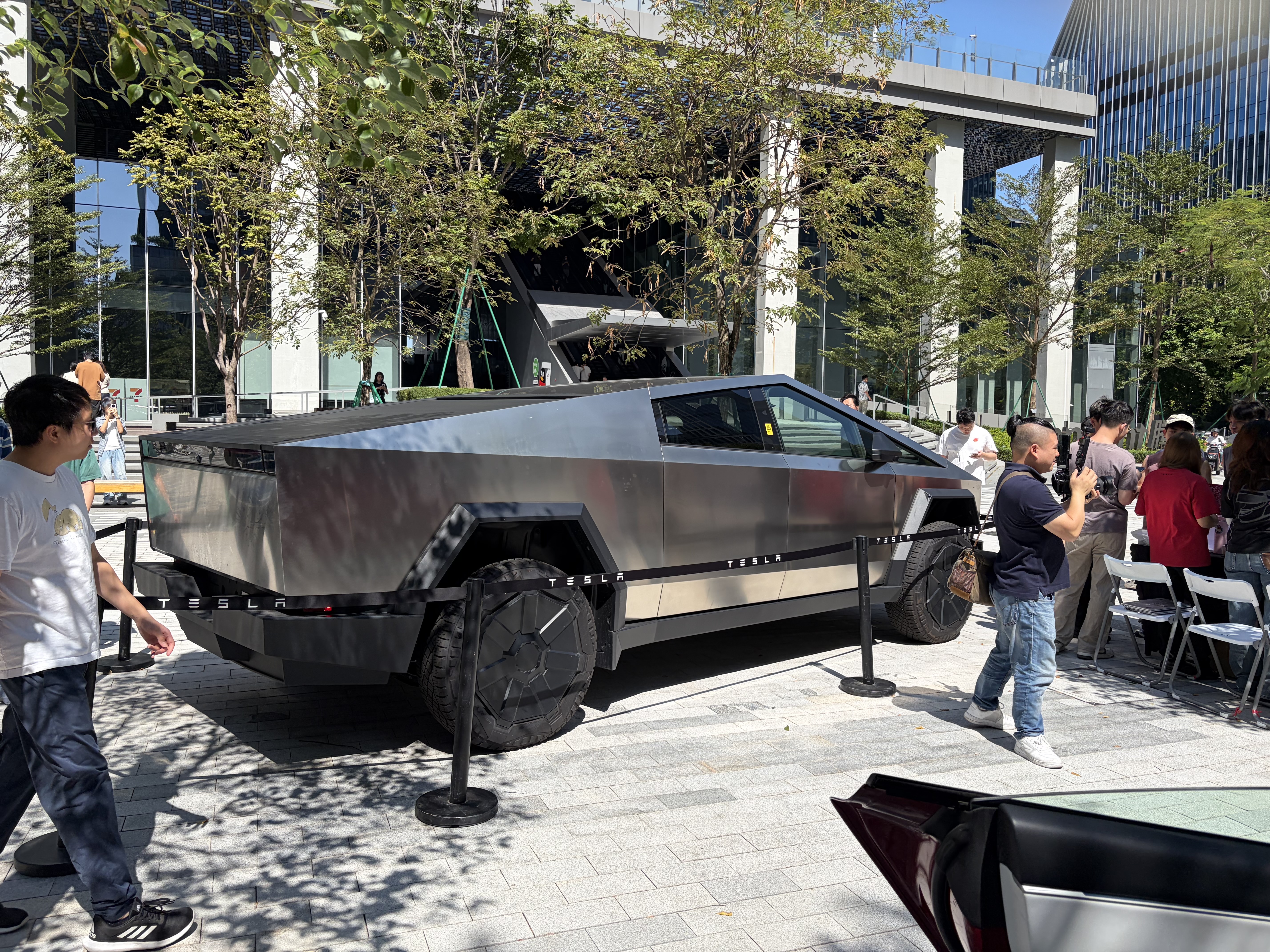 A crowd gathers around a Tesla Cybertruck displayed outdoors beneath trees in front of a modern glass-fronted building. Black barriers marked “TESLA” surround the vehicle.