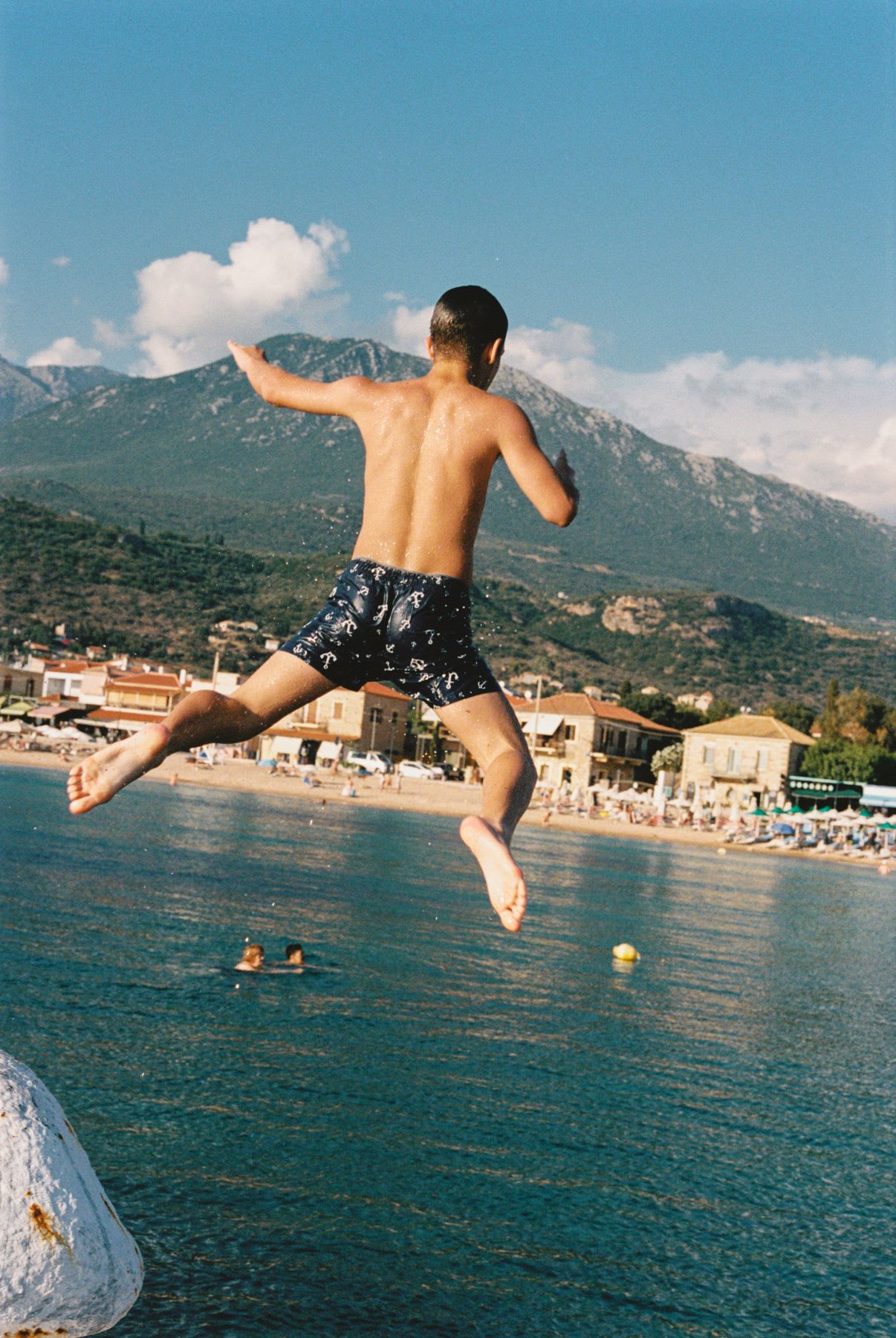 A boy in swim trunks leaps into the sea on a sunny day, with a scenic coastal town and mountains in the background.