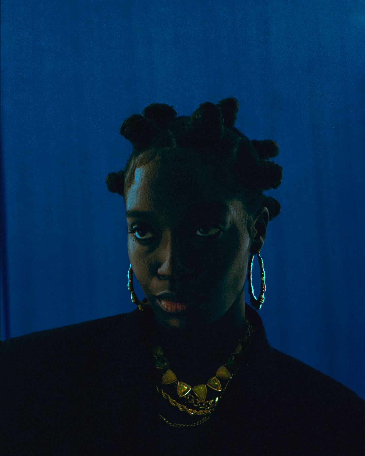 Woman wearing multiple necklaces poses against a blue background.