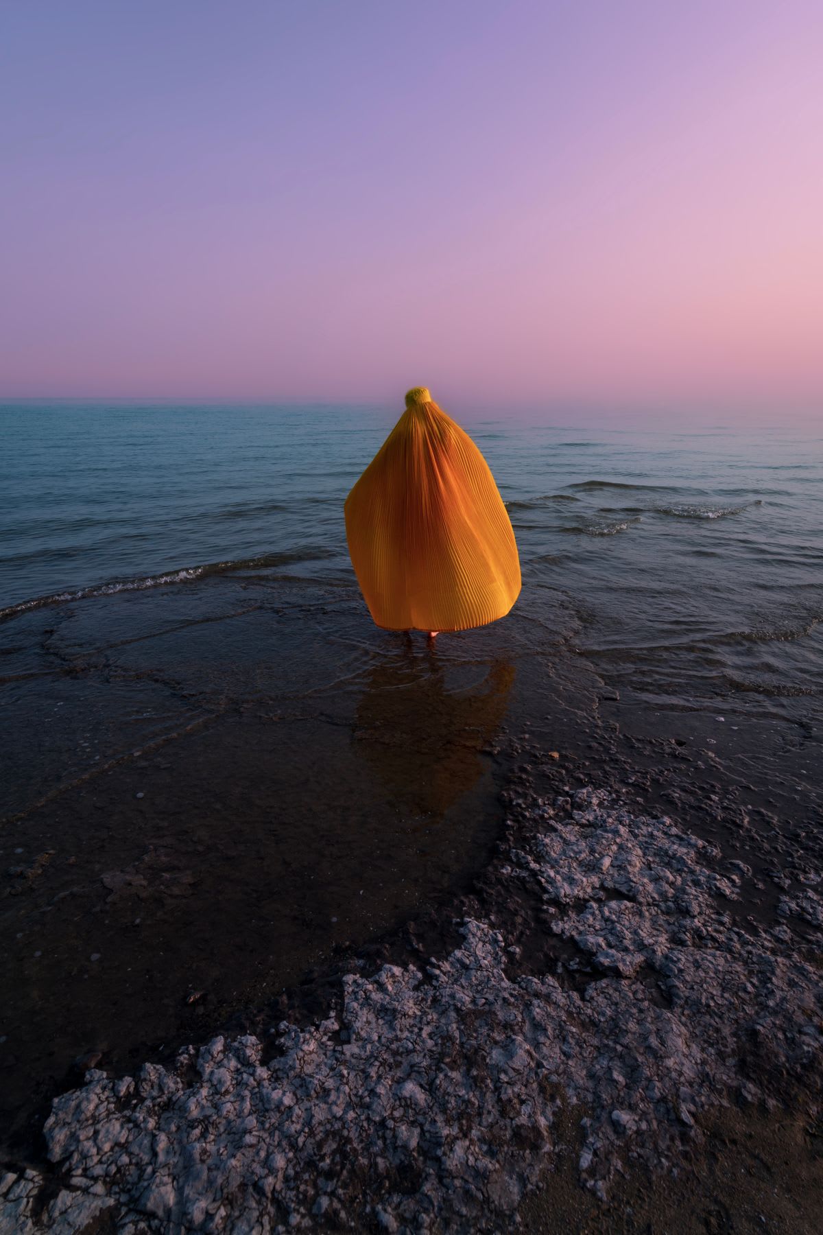 A person covered in a yellow cloth is standing on the edge of the sea at dusk, with the sky in shades of pink and purple.