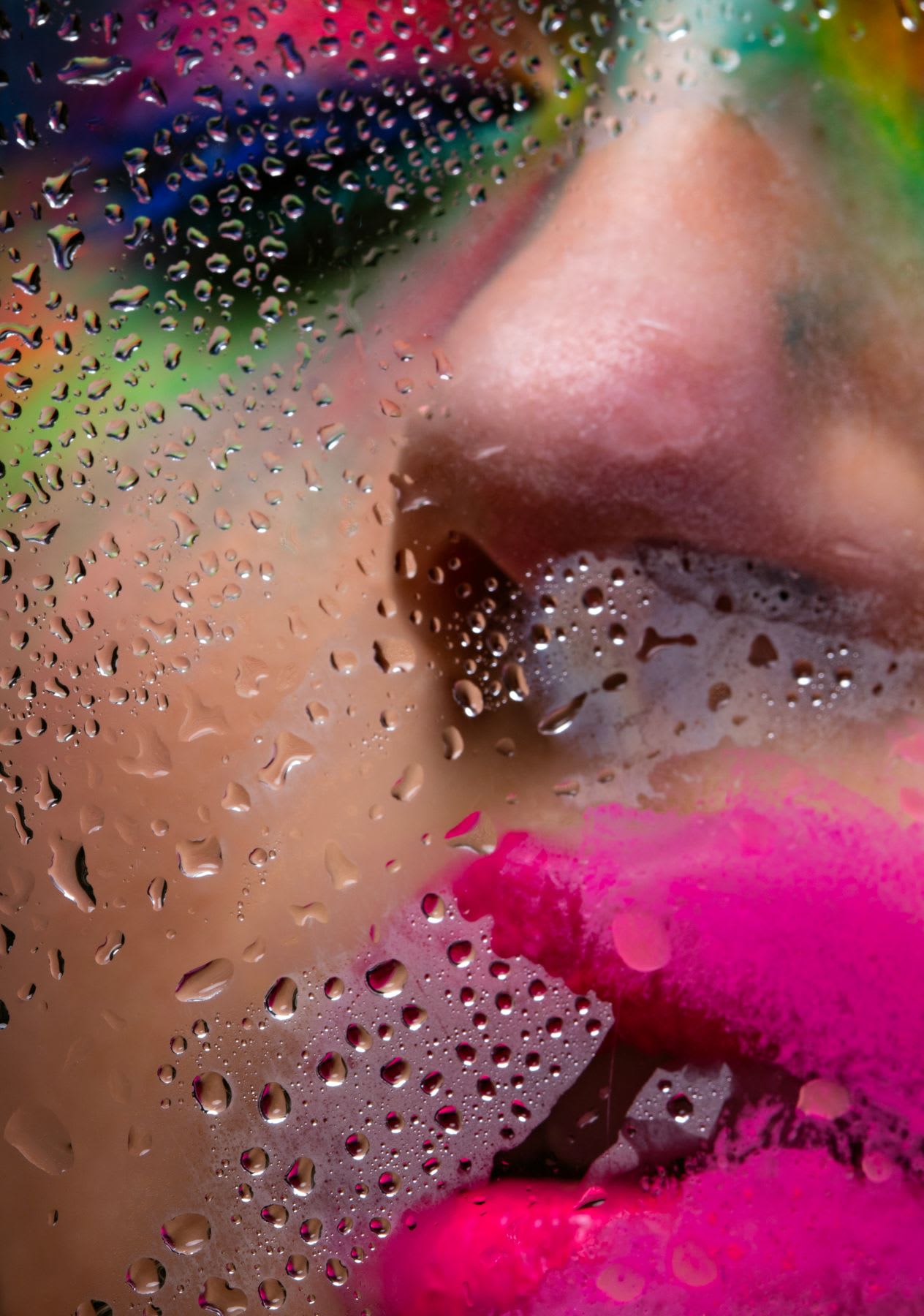 Portrait of a woman wearing bright and colorful makeup while outside with her face touching wet glass in the daytime.