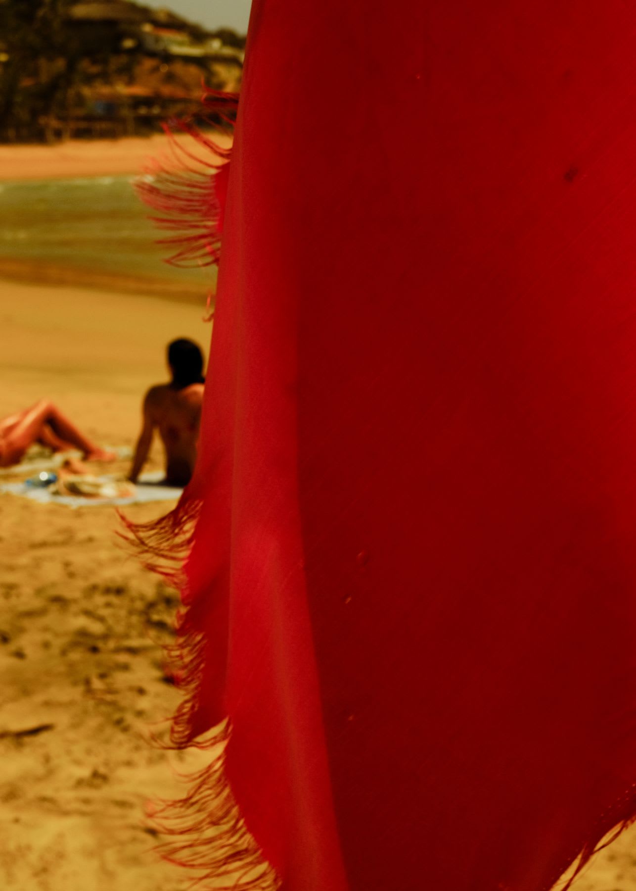A close-up of a red fringed towel on a sunny beach where people are relaxing on the sand in the background.